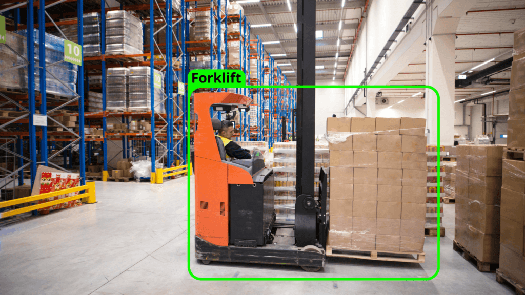 Worker driving an orange forklift in a warehouse, lifting a pallet of cardboard boxes. The warehouse has tall storage racks filled with goods and a spacious, well-lit interior.